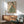 May include: Framed poster showing Japanese woman in green striped kimono with coral floral sleeves, holding persimmon fruit, displayed above wood sideboard with white ceramic table lamp in modern living room setting with sage green upholstered chair and natural lighting casting shadows on white wall