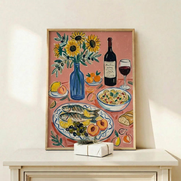 May include: Framed art print displayed on light wood console table with small white gift box. Artwork shows Mediterranean feast scene with sunflowers in blue vase, grilled fish with lemon and olives on decorative plate, pasta bowl, wine bottle and glass, fresh fruits, and bread against coral pink background. Warm, illustrative style with golden wood frame.