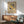 May include: Framed Bauhaus exhibition poster with geometric design featuring yellow and black circles arranged around central striped pattern on cream background, displayed above mid-century modern wood credenza with white ceramic lamp, next to sage green upholstered armchair with wooden frame in sunlit room with white walls.