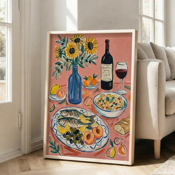 May include: Large framed poster with coral pink background showing Mediterranean feast illustration featuring sunflowers in blue vase, wine bottle and glass, grilled fish with lemon and olives on decorative plate, pasta bowl, fresh fruits, and bread. Artwork displayed in bright white room next to beige armchair near tall windows, leaning against white wall on light wooden floor.