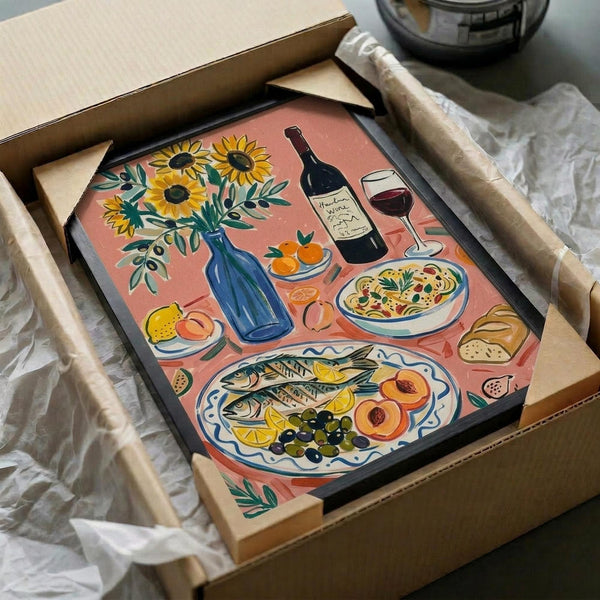 May include: Framed illustration artwork in coral pink showing Mediterranean feast with sunflowers in blue vase, wine bottle and glass, pasta bowl, fish platter with citrus and olives, fresh fruit, and bread. Black frame nestled in cardboard shipping box with brown paper wrapping on white tissue paper.