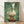 May include: Framed art print leaning against white wall on light wooden floor. The artwork shows a white duck or goose with orange beak and feet, standing on a wooden surface with a toilet paper roll balanced on its head. Painted in realistic style with muted green-blue textured background. Natural wood frame visible around the print.