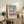 May include: Modern architectural poster in light wood frame displayed on rustic wooden sideboard. Shows pink and teal geometric building design with Bauhaus text below. Scene includes fern plant, ceramic vase, books, and brown leather chair in warm-toned living room setting with cream walls.