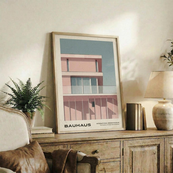 May include: Modern architectural poster in light wood frame displayed on rustic wooden sideboard. Shows pink and teal geometric building design with Bauhaus text below. Scene includes fern plant, ceramic vase, books, and brown leather chair in warm-toned living room setting with cream walls.