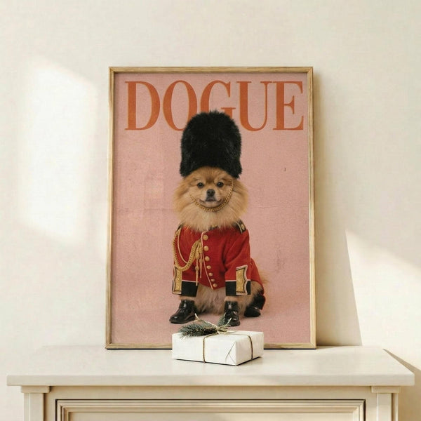 May include: Framed poster with pink background showing a fluffy Pomeranian dog wearing a red British Royal Guard uniform with gold details, black tall fuzzy hat, and small black boots. The word "DOGUE" appears in orange letters at the top. Gold frame sits on white wooden shelf next to small wrapped gift with evergreen sprig against cream wall.