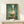 May include: Framed artwork showing a white duck with orange beak and feet balancing a toilet paper roll on its head, painted in realistic style against green textured background. Wood frame displayed on light wooden surface next to small wrapped gift with twine bow on white wall.