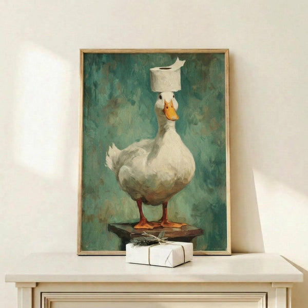 May include: Framed artwork showing a white duck with orange beak and feet balancing a toilet paper roll on its head, painted in realistic style against green textured background. Wood frame displayed on light wooden surface next to small wrapped gift with twine bow on white wall.