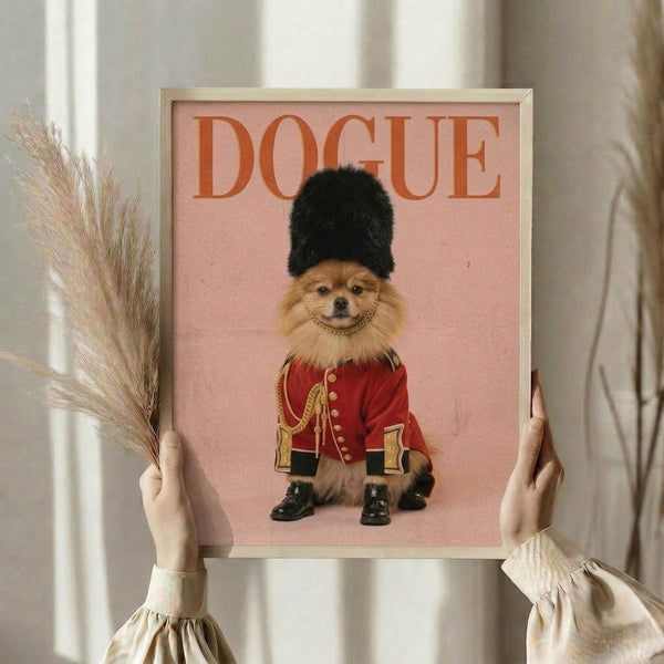 May include: Hands holding a framed poster featuring a small Pomeranian dog dressed as a British Royal Guard in red military jacket with gold buttons, black fur hat, and tiny black boots against a pink background with "DOGUE" text at top. The light wood frame is displayed against white curtains with dried wheat stems visible on the left side.