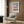 May include: Modern cream armchair with geometric patterned pillow in contemporary living room setting. Framed Bauhaus architectural poster featuring pink modernist building against blue-green background hangs on wood-paneled wall. Gold floor lamp, dark side table with open book, and neutral curtains complete the minimalist interior design scene.