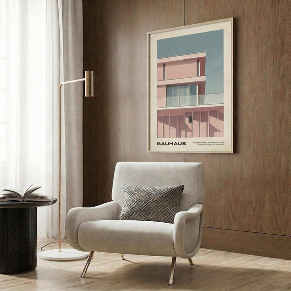 May include: Modern cream armchair with geometric patterned pillow in contemporary living room setting. Framed Bauhaus architectural poster featuring pink modernist building against blue-green background hangs on wood-paneled wall. Gold floor lamp, dark side table with open book, and neutral curtains complete the minimalist interior design scene.