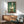 May include: A framed artwork showing a white duck balancing a toilet paper roll on its head, painted in realistic style against a teal-green textured background. The poster hangs above a mid-century modern wooden credenza with a white ceramic table lamp. A sage green upholstered armchair with wooden frame sits nearby on a round wooden coffee table, all in a bright room with white walls and natural sunlight casting shadows.