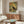 May include: Framed vintage-style poster showing black silhouette of heron against warm yellow, orange and teal brushstroke background, displayed above walnut mid-century credenza in modern living room with white curved chair, oval glass coffee table, and brass floor lamp