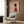 May include: A framed poster on a wood-paneled wall showing a fluffy Pomeranian dog dressed as a British Royal Guard in red military uniform with gold details, black boots, and tall black fur hat, set against a pink background with "DOGUE" text. The room features a beige upholstered chair with geometric pillow, brass floor lamp, and dark side table with open book.