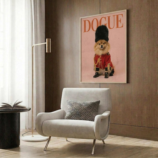 May include: A framed poster on a wood-paneled wall showing a fluffy Pomeranian dog dressed as a British Royal Guard in red military uniform with gold details, black boots, and tall black fur hat, set against a pink background with "DOGUE" text. The room features a beige upholstered chair with geometric pillow, brass floor lamp, and dark side table with open book.