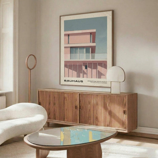 May include: Modern living room displaying framed Bauhaus poster featuring pink geometric building against blue sky. White upholstered chair, walnut credenza, oval glass coffee table, and brass floor lamp create mid-century aesthetic. Poster shows clean architectural lines with "BAUHAUS INTERNATIONAL DESIGN MUSEUM" text below building image.