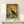 May include: Framed vintage-style poster with earthy orange, yellow, and green brushstroke background featuring black silhouette of a long-necked wading bird. Text reads "WETLAND ECOLOGY FIELD GUIDE" at top and "MID-CENTURY MARSHLAND" at bottom. Displayed on light wooden shelf with small wrapped gift box beside it against cream colored wall.