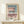 May include: Framed architectural poster with modernist building design in muted pink, sage green and white tones, displayed on light wood surface against cream wall. Small wrapped gift with evergreen sprig sits nearby. Thin wooden frame with white matting showcases geometric building facade with clean lines and minimalist aesthetic.