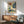 May include: Framed vintage-style botanical poster featuring colorful illustrated flowers and plants in blues, oranges, and greens with "OSAKA '82 FLOWER MARKET" text, displayed above a wooden sideboard with white ceramic lamp in modern living room setting with teal upholstered chair and wooden coffee table.