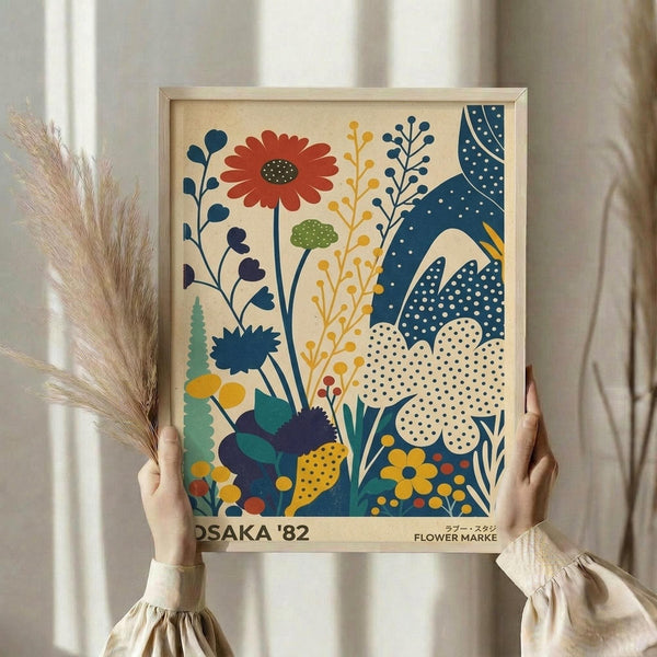 May include: Hands holding a framed botanical print featuring stylized flowers in orange, blue, yellow and green with polka dot patterns. The artwork shows various wildflowers and plants in a mid-century modern illustration style against a cream background. Natural wheat stems are visible to the left, and the scene is photographed against white curtains in soft natural lighting.