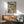 May include: Framed botanical fish print hanging above wooden sideboard in mid-century modern living room. Three speckled fish swim among green aquatic plants and coral dots on pale seafoam background. Warm wood frame matches furniture. Teal upholstered chair with wooden arms sits beside sideboard topped with white ceramic lamp. Natural sunlight casts shadows across white walls.