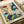 May include: Vintage-style poster print with colorful illustrated flowers and plants in orange, blue, yellow, and green. Features stylized botanical elements including daisies, leaves, and dotted mushroom shapes against cream background. Text reads "OSAKA '82" and "FLOWER MARKET" in bottom corners. Print shown on wooden surface with rolled white paper in background.