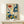 May include: Framed vintage-style botanical poster with colorful stylized flowers and plants in blues, oranges, yellows and greens on cream background, displayed on light wood shelf with small wrapped white gift box, against white wall in minimalist room setting