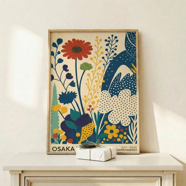 May include: Framed vintage-style botanical poster with colorful stylized flowers and plants in blues, oranges, yellows and greens on cream background, displayed on light wood shelf with small wrapped white gift box, against white wall in minimalist room setting