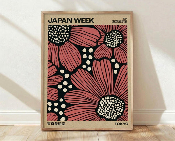 May include: Framed vintage-style poster featuring large pink and coral flowers with black centers filled with white dots, set against a black background with scattered white polka dots. The poster displays "JAPAN WEEK" text at top with Japanese characters and "1984" and "TOKYO" in corners. Natural wood frame sits on light wooden flooring against white wall with soft shadows.