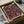 May include: Vintage exhibition poster with large pink sunflowers and black dotted stems on dark background, displayed in wooden frame inside cardboard shipping box with protective paper wrapping and wooden dowels, sitting on white fabric surface next to glass bowl.