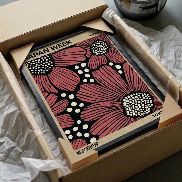 May include: Vintage exhibition poster with large pink sunflowers and black dotted stems on dark background, displayed in wooden frame inside cardboard shipping box with protective paper wrapping and wooden dowels, sitting on white fabric surface next to glass bowl.