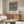 May include: Framed vintage exhibition poster featuring two colorful birds perched among strawberry plants and flowers on blue background, displayed above mid-century modern walnut credenza in minimalist living room with cream chair, glass coffee table, and neutral walls