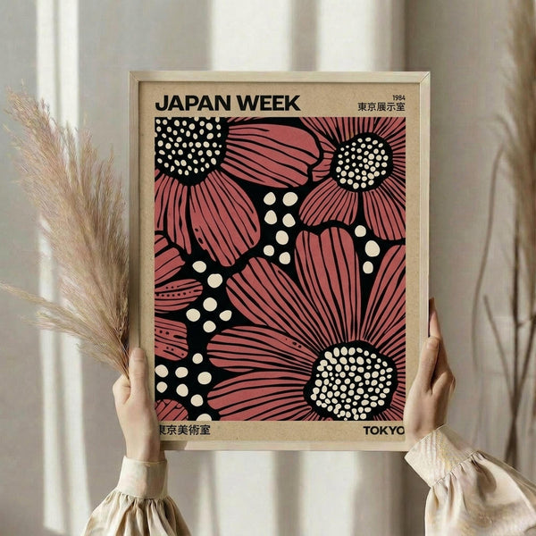 May include: Person in cream knit sweater holding a framed vintage exhibition poster featuring stylized pink and coral flowers with white polka dot centers on black background. The poster displays "JAPAN WEEK" at top with Japanese characters and "TOKYO" at bottom. Natural wood frame, soft lighting, dried wheat stems visible to left, neutral curtained background.