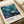 May include: Vintage-style art print displayed on cream colored paper showing an underwater scene with teal and blue fish swimming among coral and sea plants. The poster features Japanese text and "MUSEE D'ART MODERNE DE LA VILLE DE PARIS" with dates from 1984 and 1986. White ribbon visible beside the print on a light wooden surface.