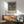 May include: Framed William Morris exhibition poster featuring two birds surrounded by strawberries and foliage in blue, red and cream colors, displayed above a wooden sideboard with white ceramic lamp in a mid-century modern room setting with sage green upholstered chair and round wooden coffee table.