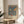 May include: Framed William Morris exhibition poster featuring two birds perched among strawberries and flowers on blue background, displayed on round wooden side table next to beige upholstered chair with decorative pillow. Natural lighting creates soft shadows on cream wall, with green plant visible in corner.