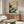 May include: Japanese-style wave artwork displayed above wooden credenza in modern living room. Print features dramatic blue-green ocean waves with white foam, flying birds, and beige sky with red text block. Warm wood furniture, white curved chair, glass coffee table, and neutral walls complete the serene interior scene.