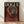 May include: Framed poster print leaning against white wall on light wood floor. Shows Rottweiler dog wearing chunky red knit sweater with hood up against dark background. Text "DOGUE" in cream letters at top mimics fashion magazine style. Natural wood frame, soft window light creating shadows on textured white wall.