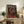 May include: Framed poster featuring a Rottweiler dog wearing a chunky red knit sweater with hood against a dark background, with "DOGUE" text at top in cream letters. The wooden-framed artwork sits on a rustic wooden sideboard alongside books, pottery, and a fern plant, with a beige upholstered chair and throw pillows visible in a warm, neutral-toned living room setting.