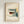 May include: Framed architectural poster featuring geometric modernist building illustration in muted tones of cream, teal, rust and sage green, showing angular structure with grid windows and stairs, displayed on light wall in wooden frame with cream matting