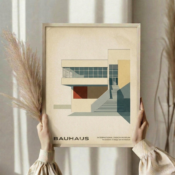 May include: Hands holding a framed architectural poster featuring a modernist building design with geometric shapes in cream, teal, orange and grey tones. The artwork shows a multi-level structure with large windows, stairs, and clean lines in Bauhaus style. Person wears a cream-colored blouse with gathered sleeves and holds dried pampas grass. Set against soft neutral curtains with natural lighting.
