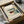 May include: Framed Bauhaus architectural print featuring geometric building design with teal grid pattern, orange and green color blocks, displayed in black frame being unwrapped from cardboard box with brown paper wrapping and white tissue paper on neutral surface.