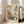 May include: Large framed botanical poster with stylized flowers in orange, teal, green and yellow colors leaning against white wall next to beige upholstered sofa. Art features folk-inspired floral designs with polka dot patterns and text reading "OSAKA '82 FLOWER MARKET" at bottom. Natural wood frame, cream colored mat, displayed on light hardwood floor in bright living room with tall windows.
