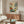May include: Framed botanical poster with colorful stylized flowers in red, blue, yellow and green displayed above a mid-century modern walnut credenza. Room features cream chair, glass coffee table with ceramic vase, and floor lamp. Poster shows folk art style wildflowers and plants with "OSAKA '82" and "FLOWER MARKET" text at bottom in wooden frame.