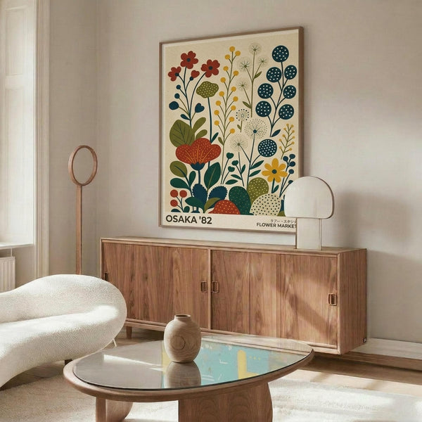 May include: Framed botanical poster with colorful stylized flowers in red, blue, yellow and green displayed above a mid-century modern walnut credenza. Room features cream chair, glass coffee table with ceramic vase, and floor lamp. Poster shows folk art style wildflowers and plants with "OSAKA '82" and "FLOWER MARKET" text at bottom in wooden frame.