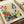 May include: A vintage-style botanical poster print featuring stylized flowers and plants in retro colors of teal, orange, olive green, and cream. The illustration shows various wildflowers, dandelions, berries, and foliage in a folk art style with dotted patterns and geometric shapes. Text reads "OSAKA '82 FLOWER MARKET" at the bottom. The print appears framed and is photographed against neutral paper wrapping materials.