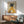 May include: Framed artwork showing a black greyhound wearing a mustard yellow turtleneck sweater and holding a white coffee cup, displayed above a wooden sideboard in a modern living room with mint green mid-century chair, white table lamp, and natural sunlight casting shadows on white walls.