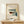 May include: Framed architectural poster featuring modernist building with geometric shapes in muted tones of teal, orange, beige and gray, displayed on light wood surface next to small wrapped gift with pine sprig, against cream wall with natural lighting