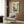 May include: Framed botanical poster with stylized flowers and plants in orange, blue, green, and yellow colors mounted on wood-paneled wall above cream upholstered armchair with geometric patterned pillow, brass floor lamp, and dark round side table with open book in modern living room setting