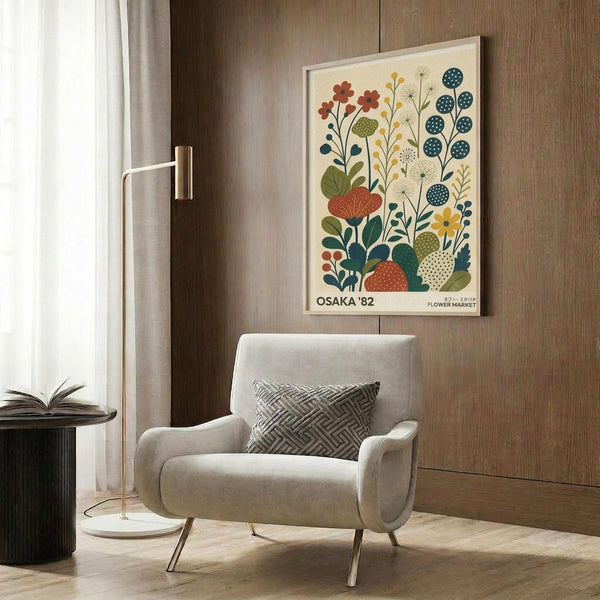 May include: Framed botanical poster with stylized flowers and plants in orange, blue, green, and yellow colors mounted on wood-paneled wall above cream upholstered armchair with geometric patterned pillow, brass floor lamp, and dark round side table with open book in modern living room setting