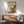 May include: Japanese woodblock print artwork displayed in modern living room with mid-century furniture. The framed poster shows a brown bird perched on orange trumpet vine flowers with mountains and traditional architecture in background. Warm peachy sky with Japanese calligraphy. Wooden sideboard below holds white ceramic lamp, sage green upholstered chair with wooden arms nearby, natural sunlight casting shadows on white wall.